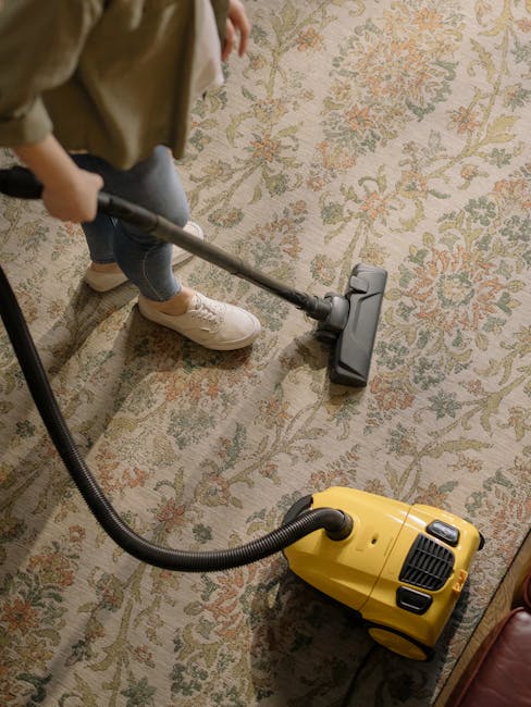 A person kneeling on a patterned carpet in a living room, using a yellow and black portable vacuum cleaner for surface cleaning. The individual is wearing a khaki shirt and blue jeans, with their hand