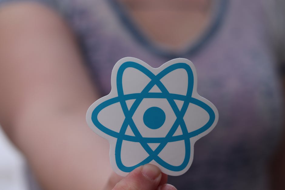 Close-up of a person holding a circular sticker with a blue atom symbol, depicting interconnected orbits and a central nucleus, in front of a blurred background of a person wearing a grey and purple t