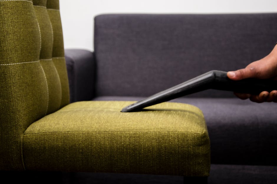 A person wearing black gloves is using a handheld electric upholstery cleaner on a dark gray fabric sofa in a living room. The sofa has a modern design with clean lines and is situated on a polished w